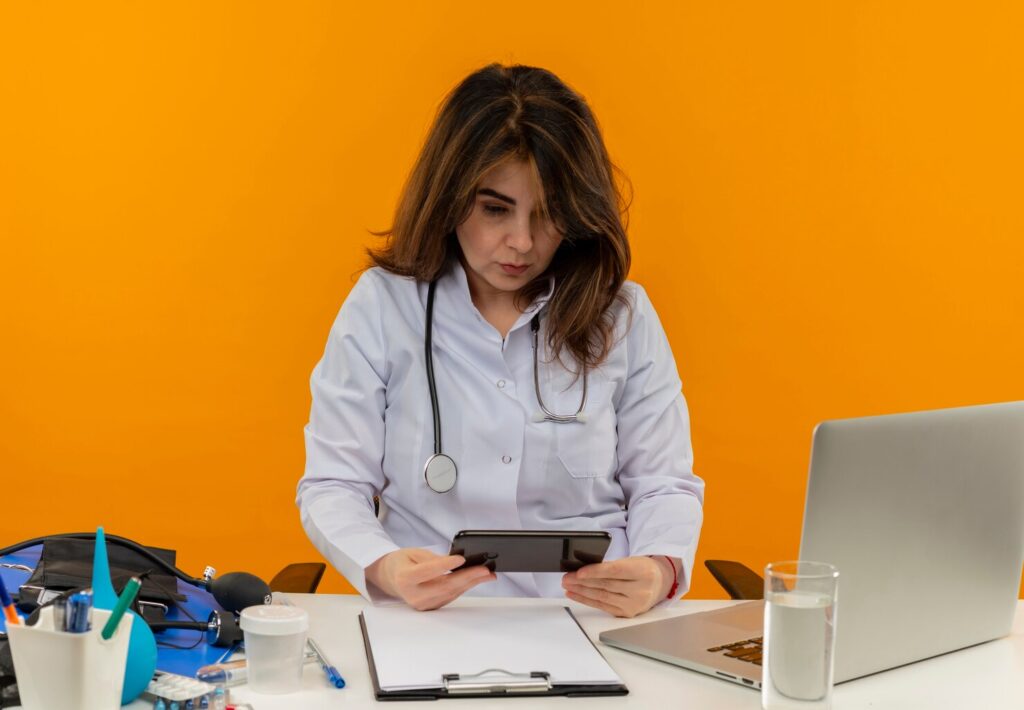 concentrated-middle-aged-female-doctor-wearing-medical-robe-stethoscope-sitting-desk-with-medical-tools-clipboard-laptop-holding-looking-mobile-phone-isolated_141793-69112 - Seahawk